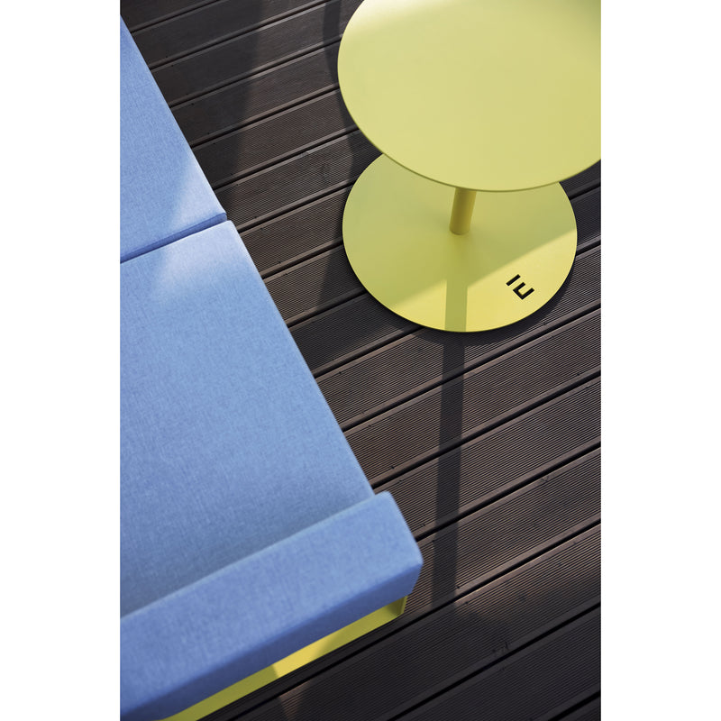 A minimalist outdoor scene displays a light blue upholstered bench and Egoes Spulka Table, a small round yellow piece with an all-metal body, set on dark wooden decking. Sunlight casts geometric shadow patterns across the surfaces.