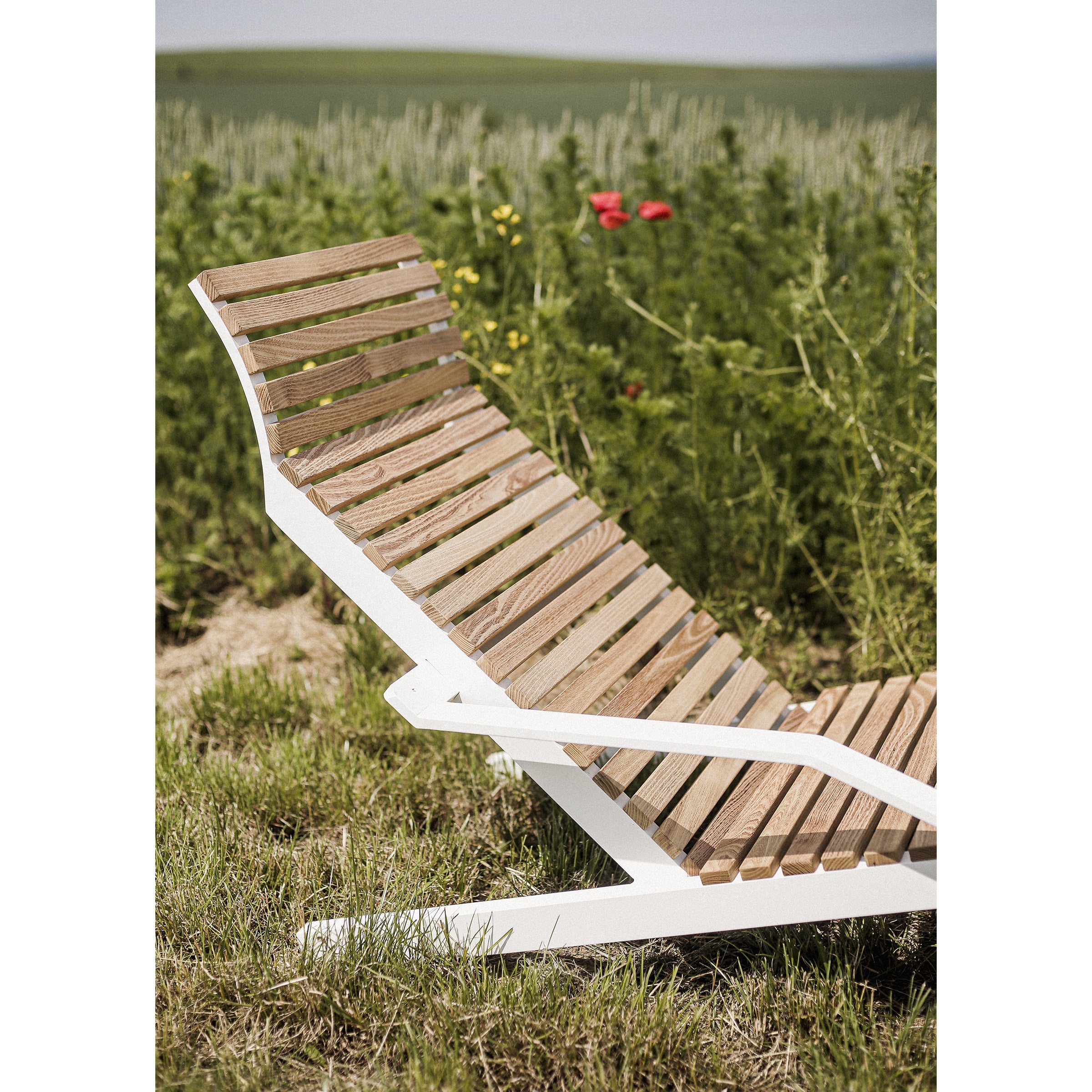 The Alva Deck Chair by Egoe, featuring a white frame, is set in a grassy field with wildflowers, rolling hills, and a blue sky. Its slatted design ensures style and lightweight durability for easy transport.