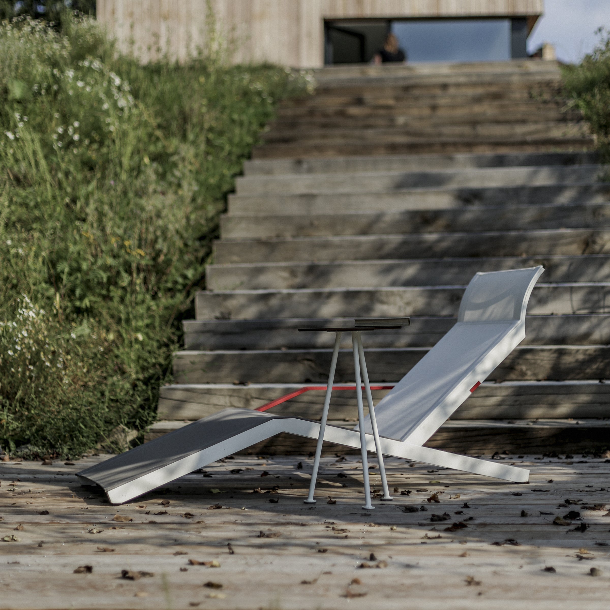 The Alva Deck Chair by Egoe, featuring a white design with a red cushion and made from durable material, sits on a wooden deck next to a small round table. Steps lead to an entrance surrounded by greenery, with leaves scattered across the deck.