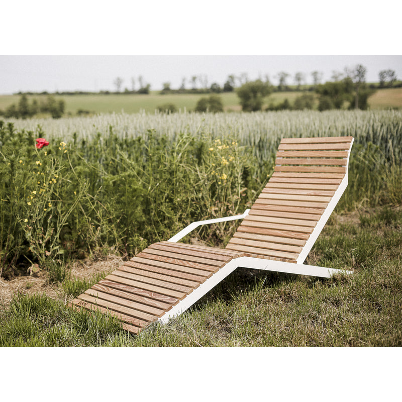An Egoe Alva Deck Chair, celebrated for its durable material and lightweight build, is elegantly positioned on grassy turf beside a field of tall green plants under a cloudy sky, with distant trees gracing the horizon.