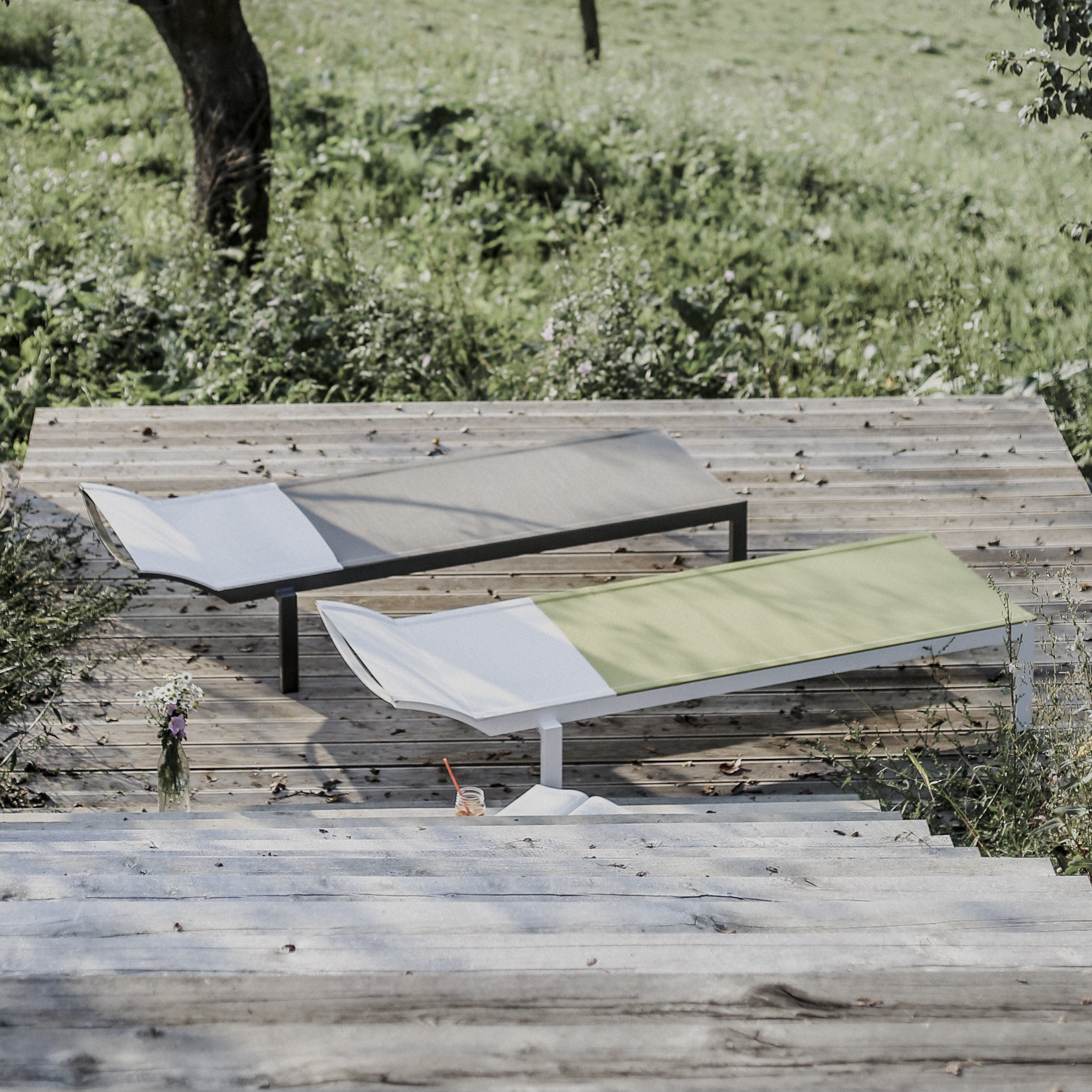 Two Alva Daybeds by Egoe, one light grey and the other light green, enhance a wooden deck amidst greenery. A small glass bottle with wildflowers adds to the serene ambiance. These stylish outdoor pieces are designed for lightweight construction, ensuring both elegance and comfort.