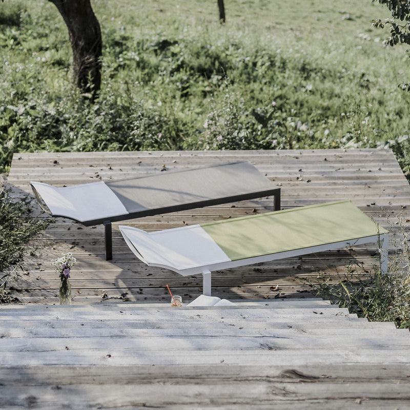 Two Alva Daybeds by Egoe, one light grey and the other light green, enhance a wooden deck amidst greenery. A small glass bottle with wildflowers adds to the serene ambiance. These stylish outdoor pieces are designed for lightweight construction, ensuring both elegance and comfort.