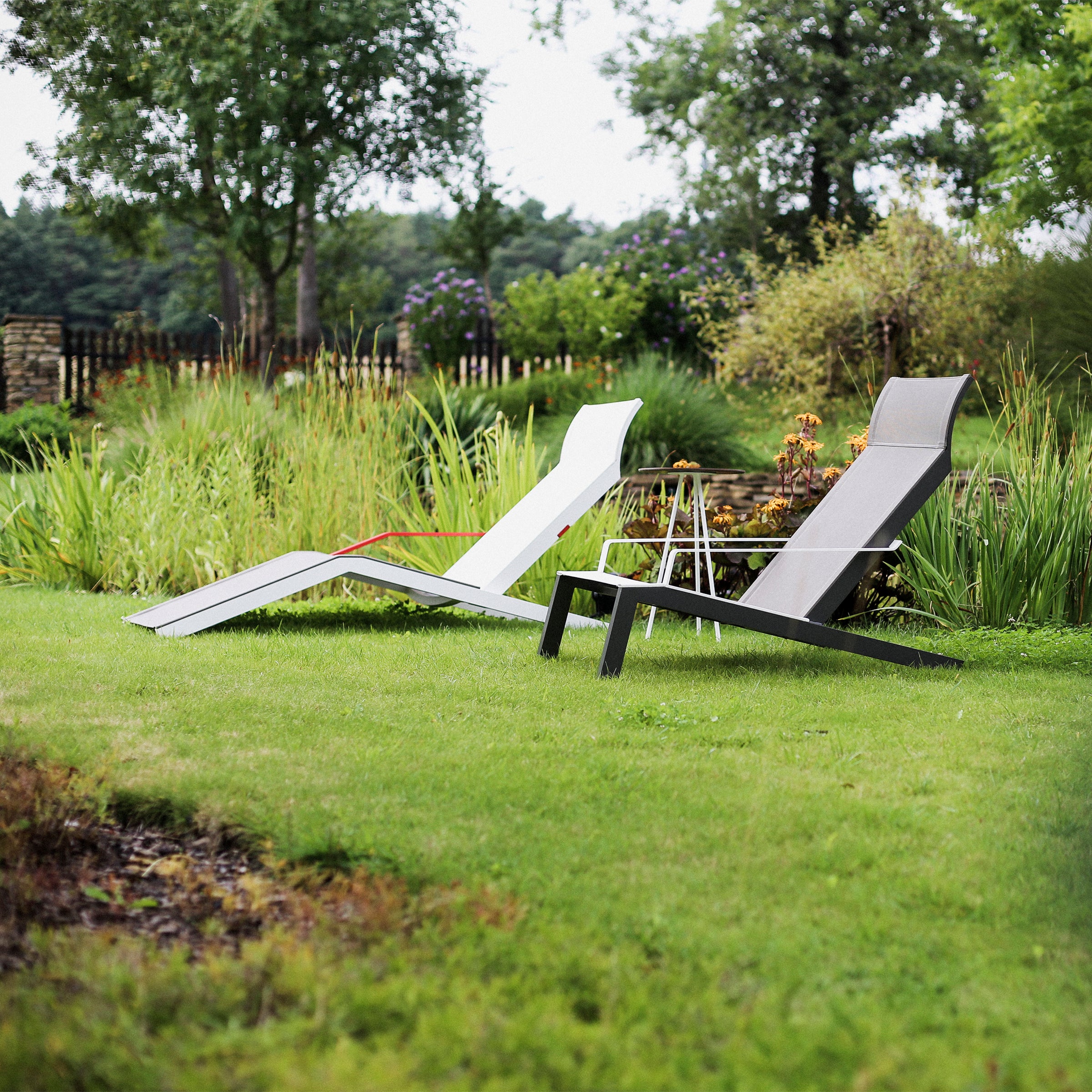 On the manicured lawn, two Alva Deck Chairs by Egoe, in white and gray, add a contemporary touch among lush greenery and a wooden fence, creating a peaceful outdoor vibe.