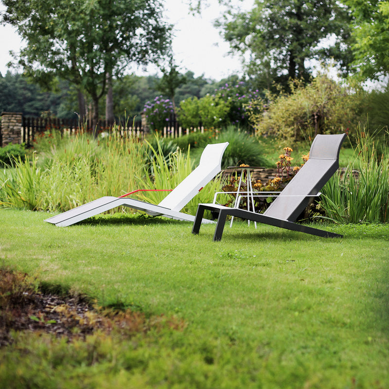 On the manicured lawn, two Alva Deck Chairs by Egoe, in white and gray, add a contemporary touch among lush greenery and a wooden fence, creating a peaceful outdoor vibe.