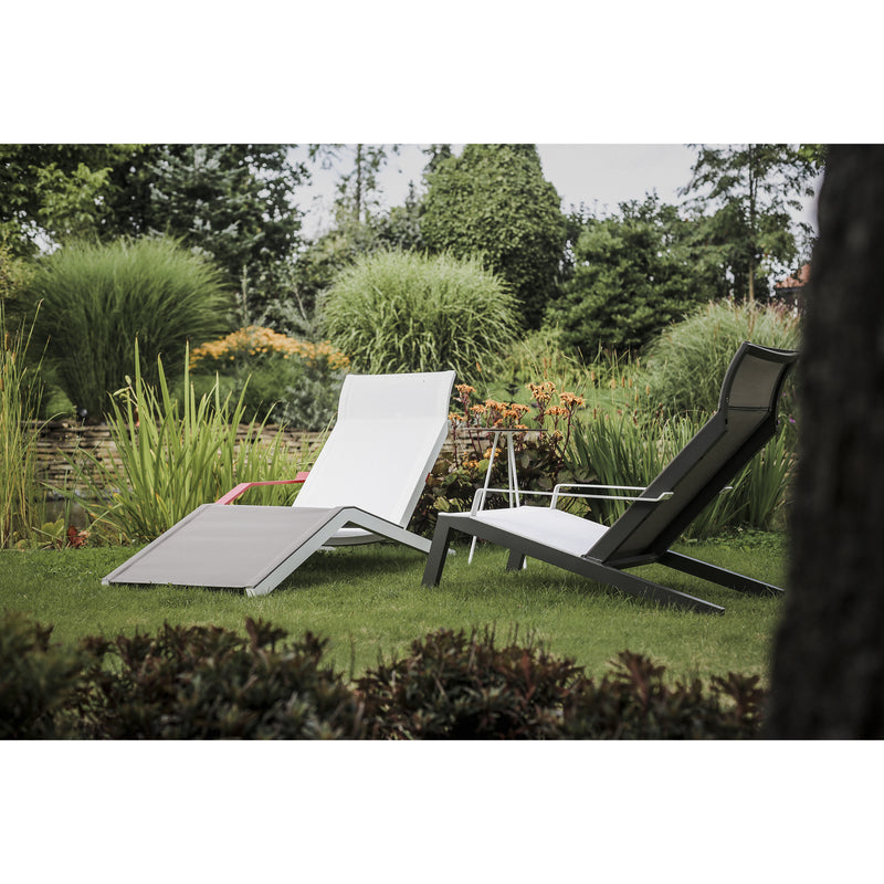 Two Egoe Alva Deck Chairs recline gracefully amid lush greenery and ornamental grasses, their lightweight construction and durable material blending seamlessly into the tranquil garden setting under a clear sky.