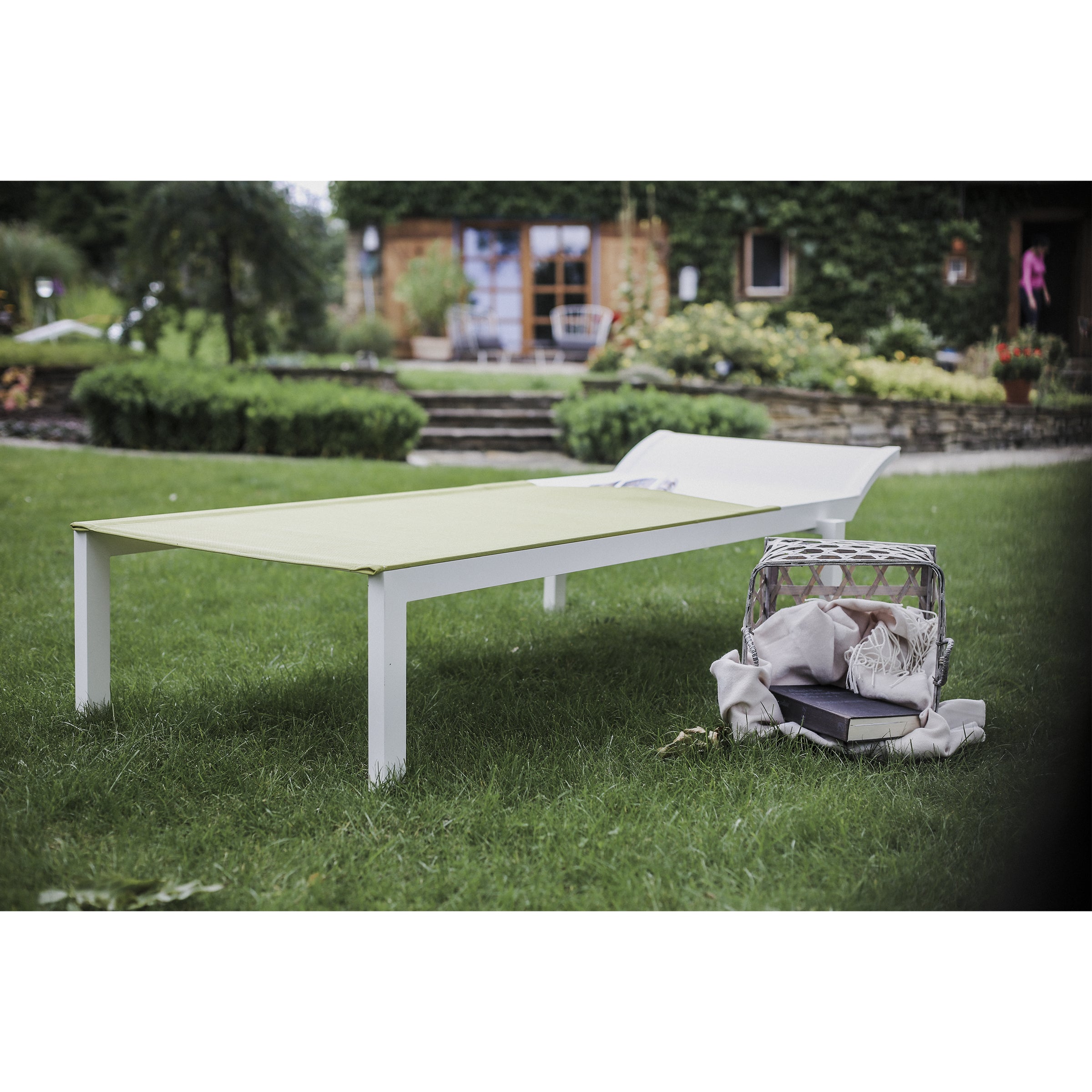 A white Alva Daybed by Egoe, featuring a green cushion, adorns the lawn, highlighting its lightweight design. A beige backpack lies nearby on the grass. Lush greenery and a stone facade house with colorful flowers frame this tranquil outdoor setting.