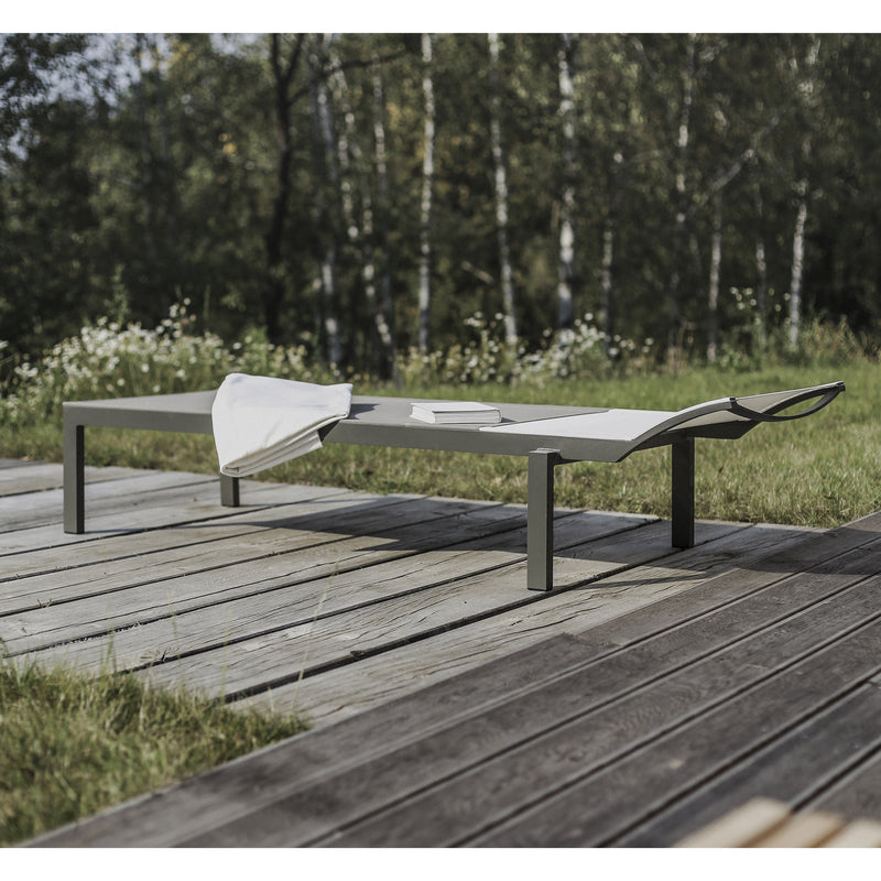 On a wooden deck, the Alva Daybed by Egoe displays its minimalist and lightweight design, adorned with a towel and book. It sits against a lush grassy area and tree grove, offering a serene setting ideal for enjoying your favorite outdoor furniture piece.