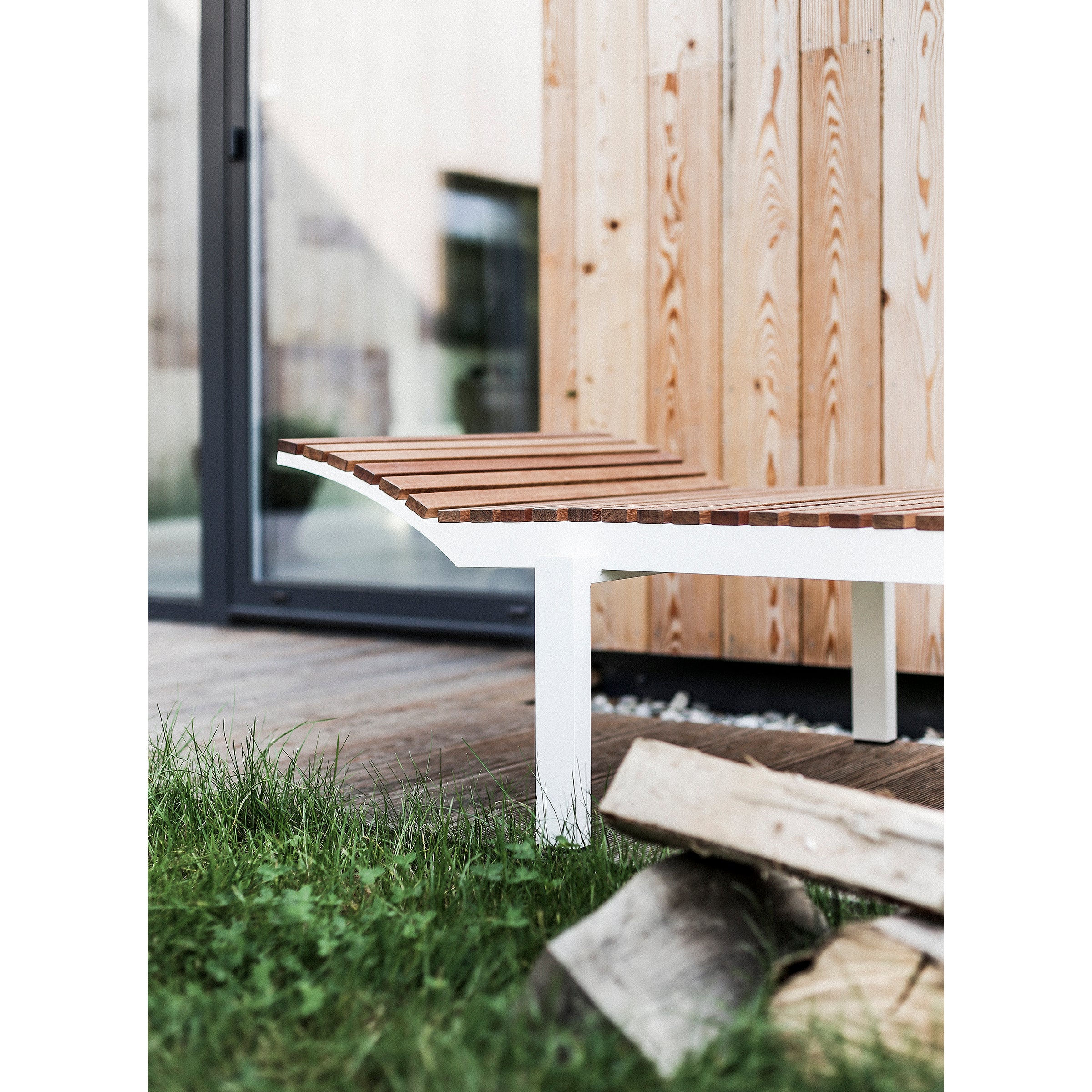 The Alva Daybed by Egoe, a modern wooden bench with a slatted seat, complements the lightweight outdoor furniture on the deck. In the background are a light wood-paneled wall and glass door, while cut firewood is stacked on the grass beside it.