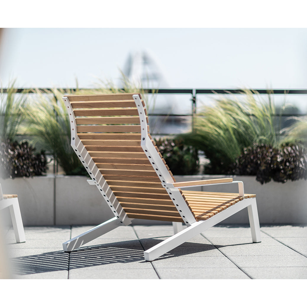 The Alva Lounge Chair by Egoe, featuring wooden slats and a white metal frame, provides lightweight comfort on a rooftop terrace. Surrounded by tall grasses and potted plants, it creates an oasis with a blurred cityscape beyond under the sunny sky.