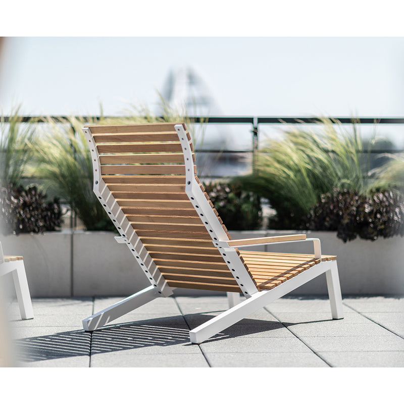 The Alva Lounge Chair by Egoe, featuring wooden slats and a white metal frame, provides lightweight comfort on a rooftop terrace. Surrounded by tall grasses and potted plants, it creates an oasis with a blurred cityscape beyond under the sunny sky.