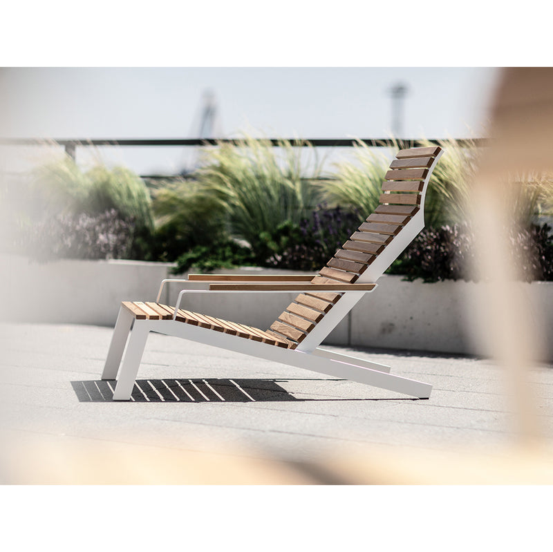 The Alva Lounge Chair by Egoe enhances the outdoor patio with its sleek modern design, combining natural and industrial elements. Its durable wooden and metal construction blends effortlessly against blurred greenery and a hazy sky.