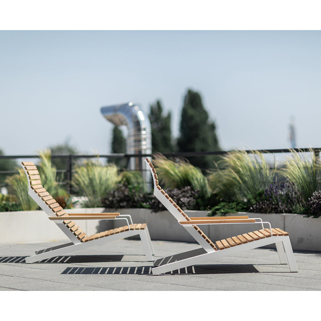 Two Alva Lounge Chairs by Egoe, with a lightweight build and wooden slats on metal frames, sit elegantly on a rooftop terrace. Tall grasses and a vent pipe form the background, underlined by a clear blue sky that completes this peaceful outdoor setting.