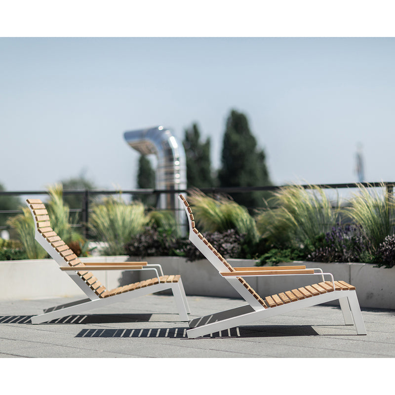 Two Alva Lounge Chairs by Egoe, with a lightweight build and wooden slats on metal frames, sit elegantly on a rooftop terrace. Tall grasses and a vent pipe form the background, underlined by a clear blue sky that completes this peaceful outdoor setting.