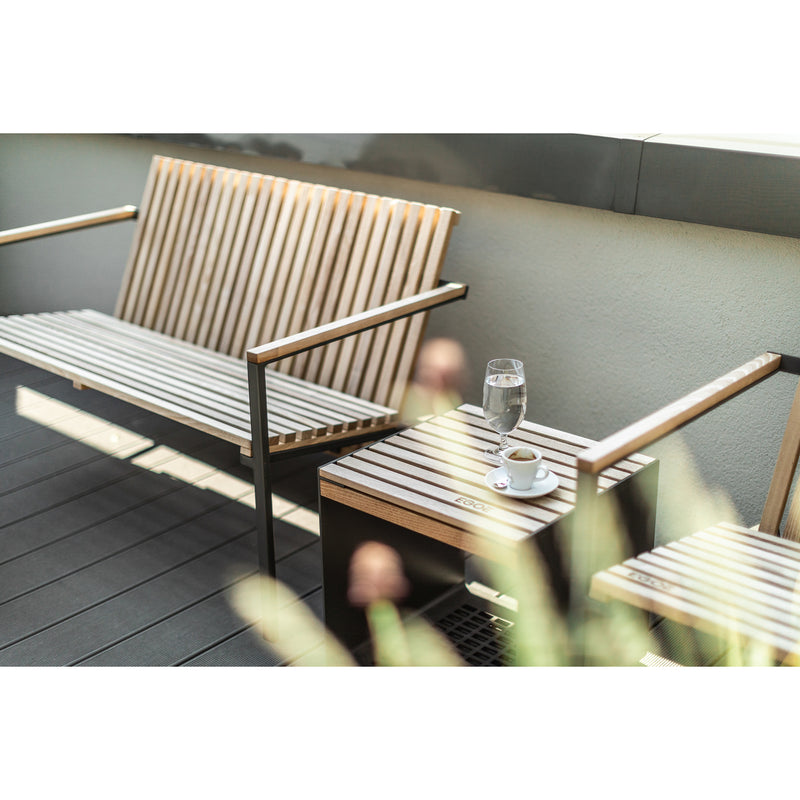 An Axis Lounge Armchair from Egoe is on a sunlit balcony next to a tropical wood table holding a glass of water and coffee. Blurred foreground plants add depth, and sunlight dances across the zinc-plated steel frame.