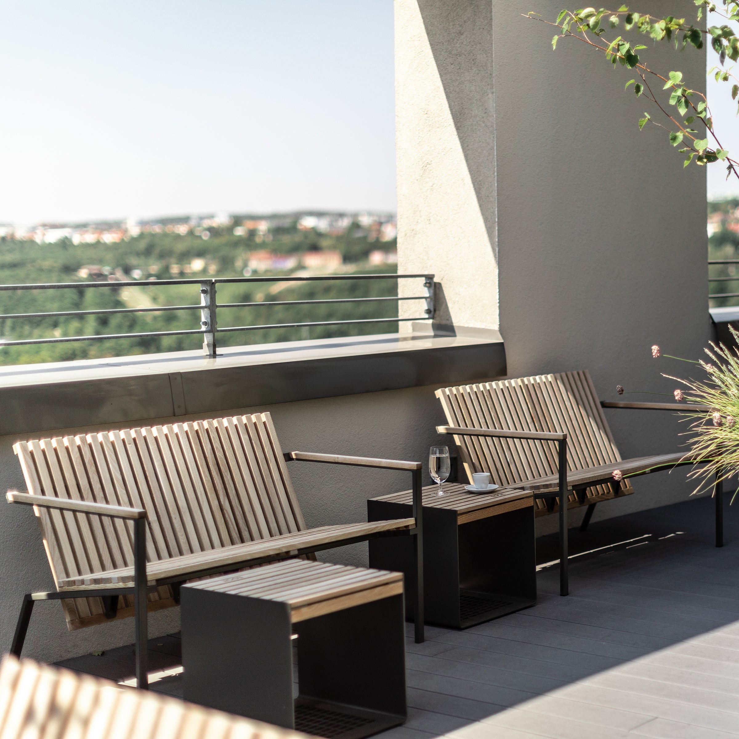 On the rooftop patio, Egoes Axis Lounge Armchairs sit beside small tables. A refreshing drink and a book rest on one table, with a zinc-plated steel railing offering views of the distant cityscape and greenery, as a tree branch gently sways in the breeze.