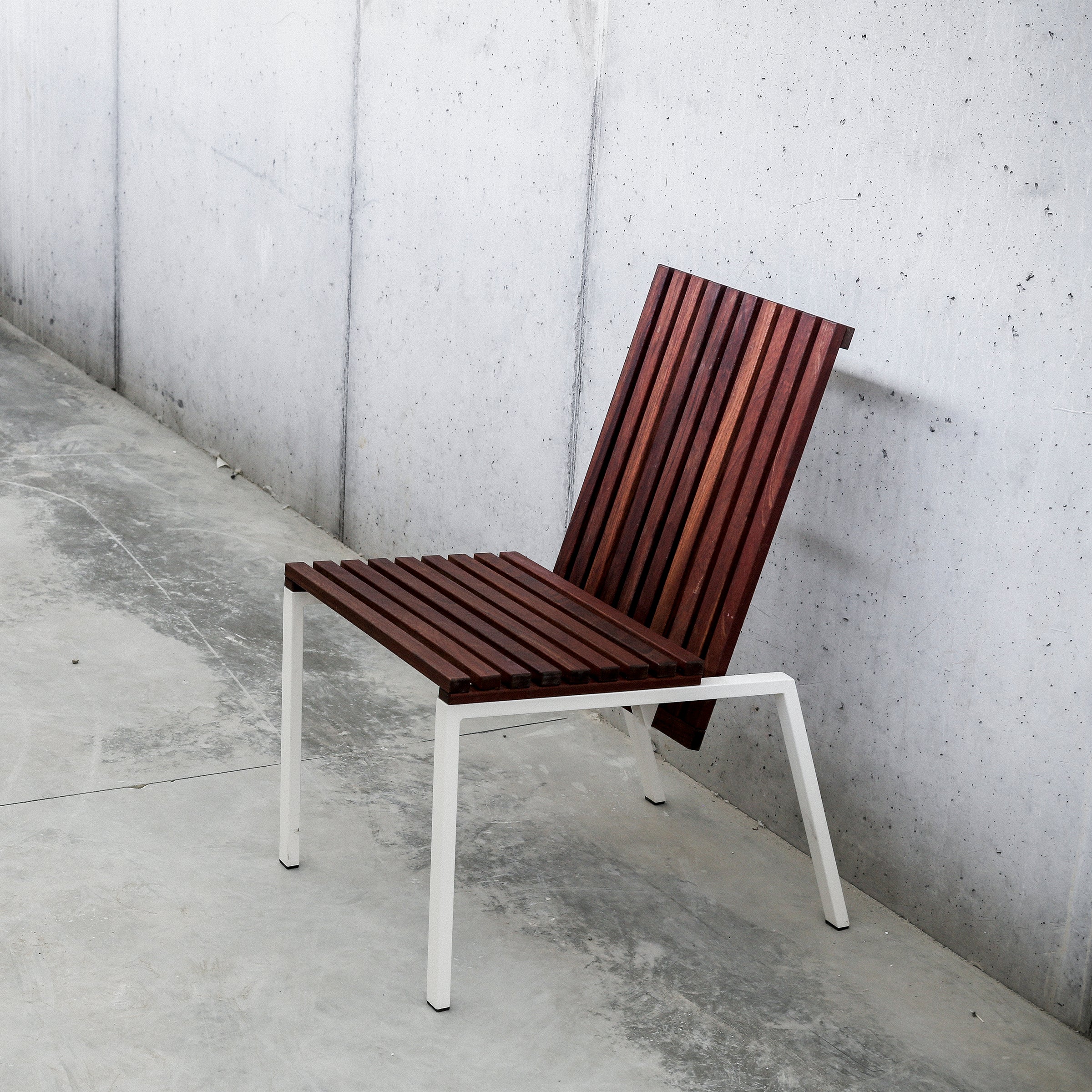Part of the Axis series by Egoe, the Axis Chair features a seat and backrest crafted from tropical wood, complemented by a sleek white zinc-plated steel frame that stands confidently on a smooth concrete floor against a grey concrete wall.
