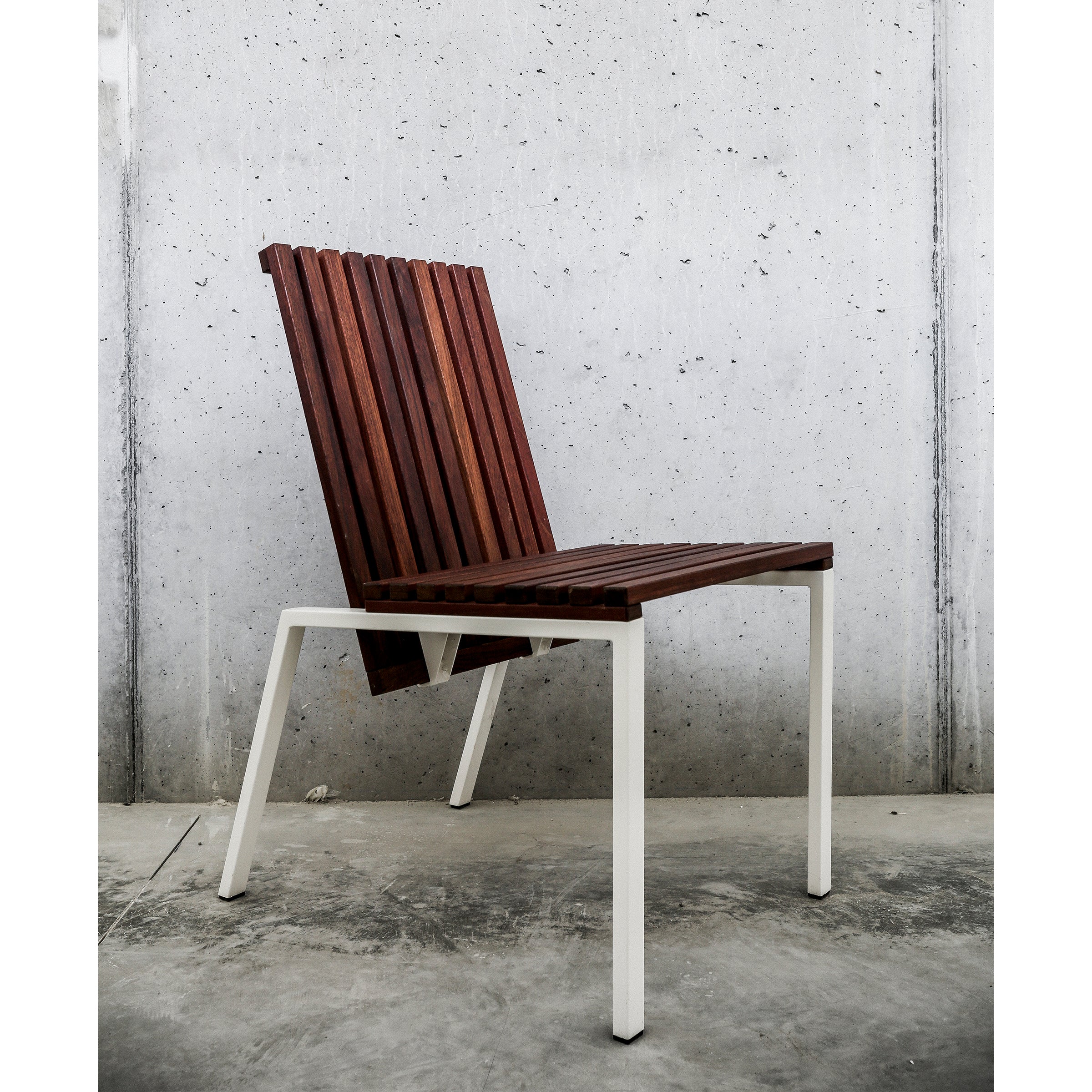 The Axis Chair by Egoe showcases a slatted tropical wood seat and backrest with white zinc-plated steel legs, set against a textured concrete wall on a concrete floor to highlight minimalist design.