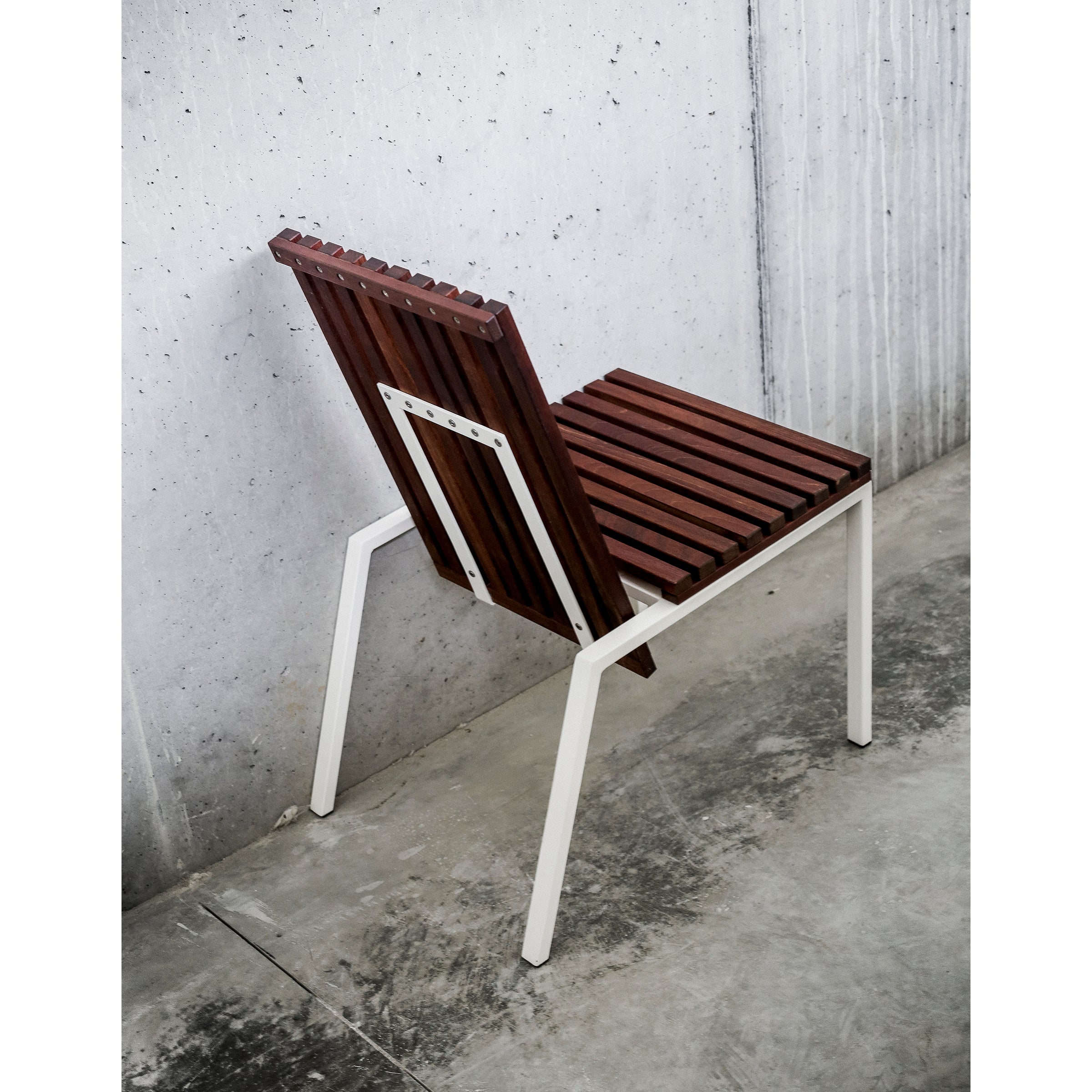 The Axis Chair by Egoe features a slatted tropical wood seat and backrest set on a sleek zinc-plated steel frame, resting on a concrete floor against a concrete wall, showcasing minimalist and industrial design.