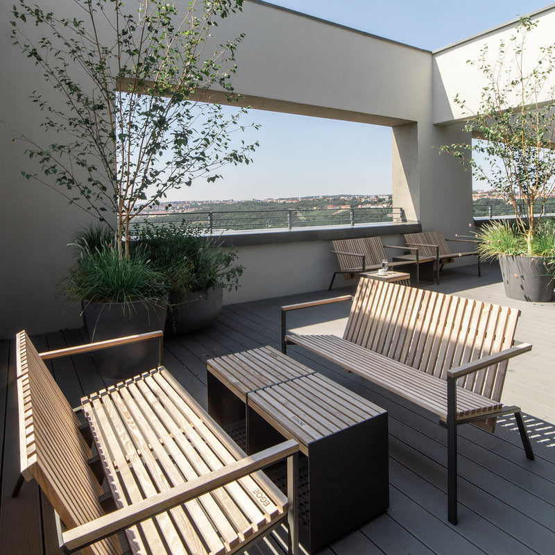 The rooftop terrace showcases benches and tables crafted from tropical wood, surrounded by large planters with lush greenery. It overlooks a cityscape framed by modern architecture under a clear blue sky. Part of Egoes Axis series, it complements the Axis Lounge Armchair perfectly.