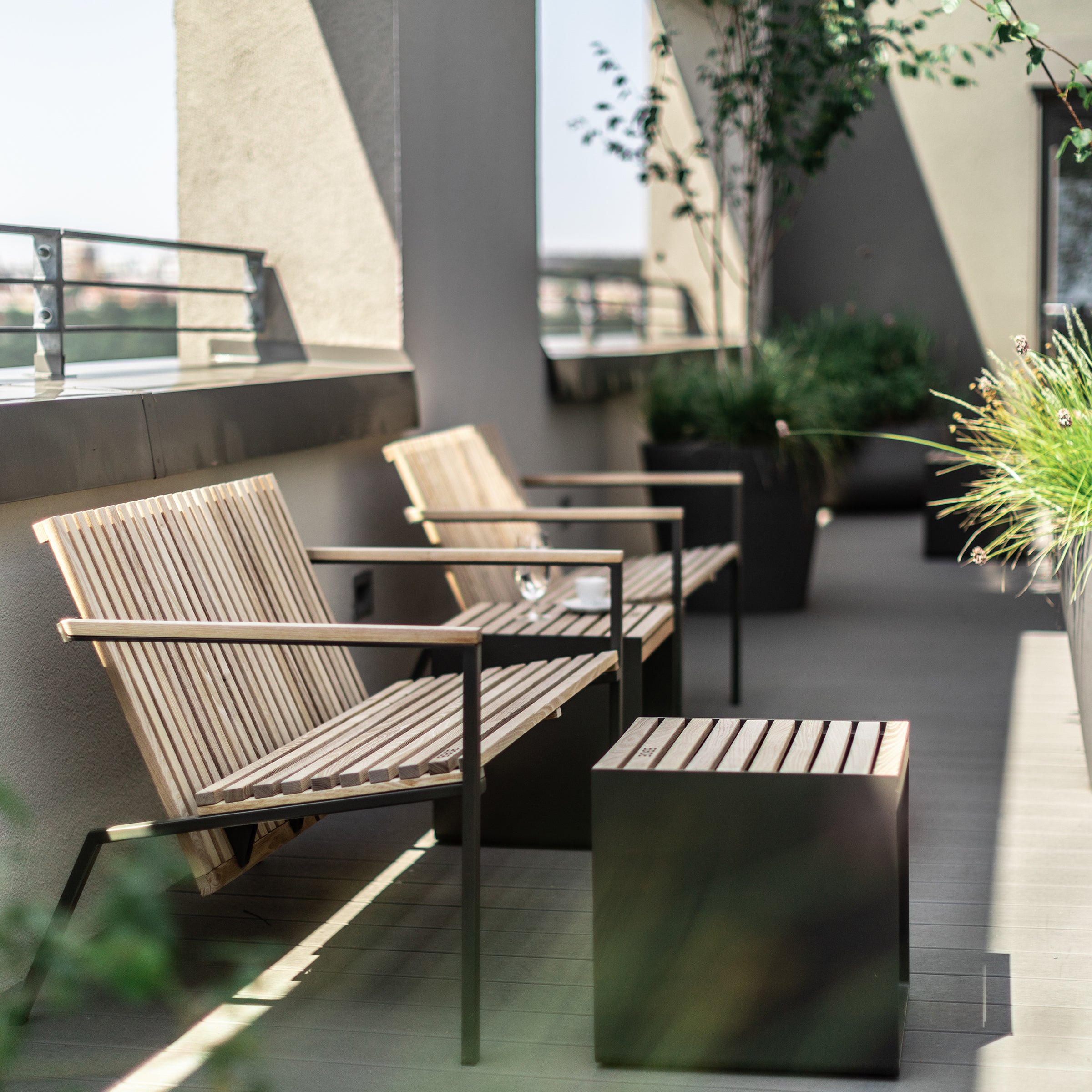 A stylish rooftop patio features Axis Lounge Armchairs by Egoe around a small table. Green plants in black pots add a natural touch as sunlight softly casts shadows, creating a calm atmosphere. The tropical wood and sleek zinc-plated steel accents enhance the design.