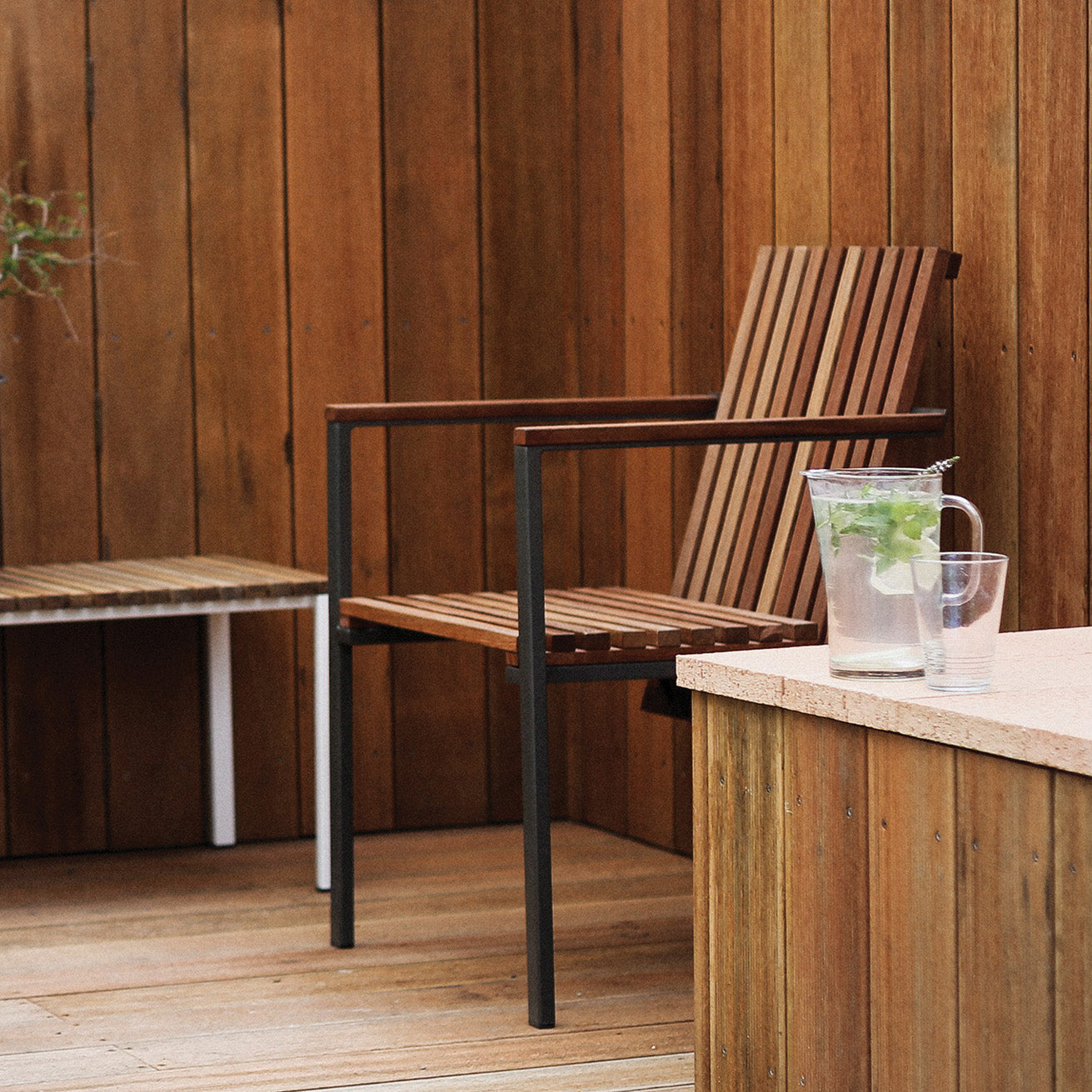 A wooden patio showcases Egoes Axis Chair and table. A mint-infused water pitcher and glass rest on the table, surrounded by tropical wood vertical panels that enhance the serene, natural outdoor ambiance.