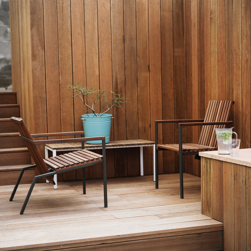 Two Axis Chairs by Egoe, featuring zinc-plated steel frames, are placed on a wooden deck. A bench between them holds a blue planter with a plant. A glass pitcher with mint-infused water sits on a ledge. Vertical wooden paneling in the background completes the serene ambiance of this Axis series setup.