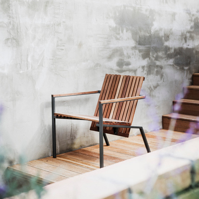 An Egoe Axis Lounge Armchair, featuring a wooden seat and zinc-plated steel frame, rests on a wooden deck. Behind it, earthy textures adorn the wall, while a staircase to the right enhances the scenes minimalistic charm.