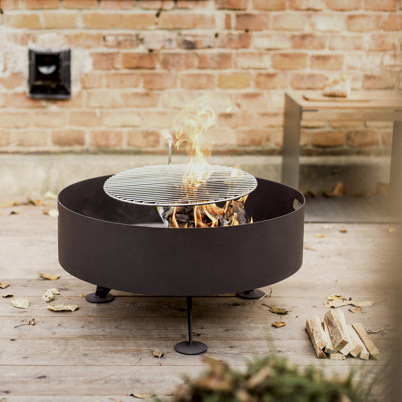 The sleek black, circular Egoe Back to Fire Fire Pit, featuring a stainless steel grill and fire-resistant color finish, burns wood amid scattered autumn leaves on a wooden deck. A brick wall and wooden bench enhance the cozy setting for grills and fireplaces enthusiasts.