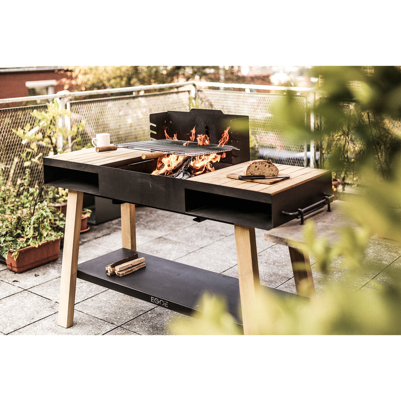 The Egoe Back to Fire Grill, featuring wooden accents, enhances patio cooking. Flames flicker as a loaf of bread sits close by, surrounded by potted plants and a metal fence, providing a view of nearby rooftops.