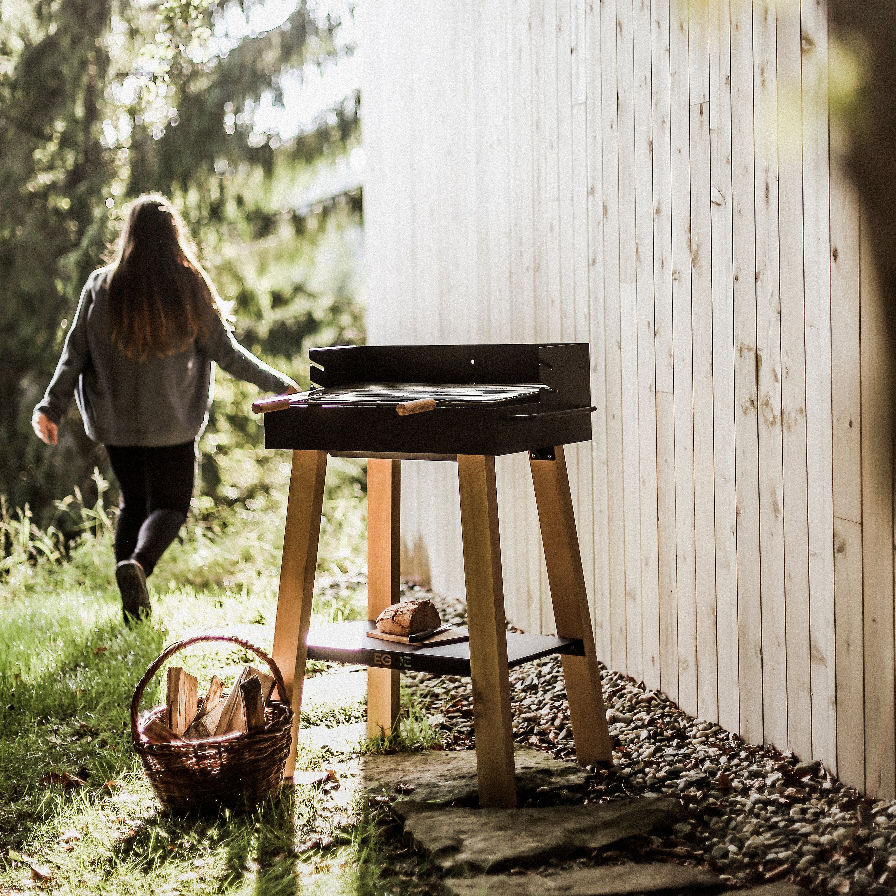 A woman walks on grass toward trees by a wooden wall. An Egoe Back to Fire Grill, with cooking utensils, sits beside a stone path. Nearby, a basket of firewood lies on the ground as sunlight filters through the trees, creating a warm ambiance.