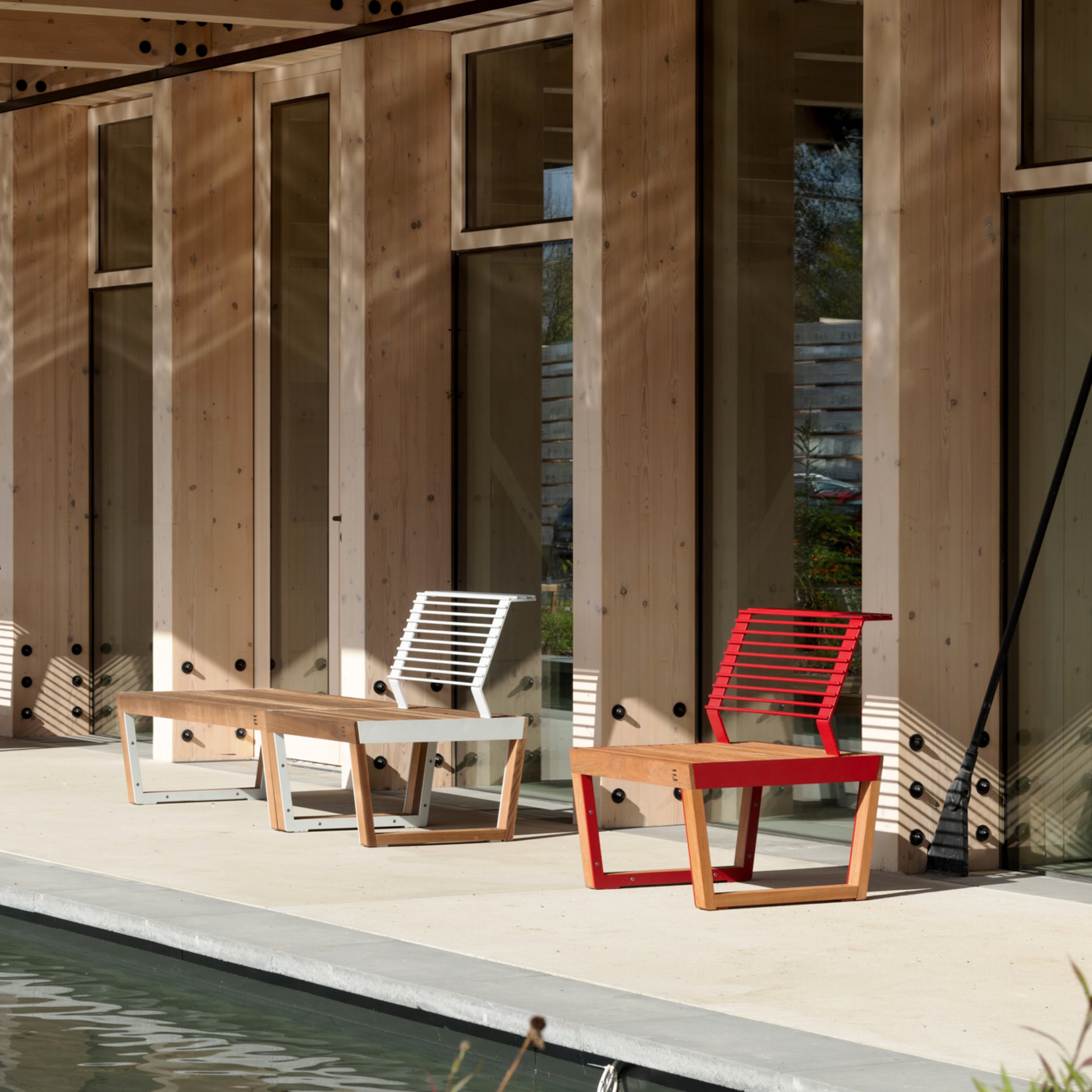 Two Barka Public Chairs from Egoe, in white and red, sit on a patio with a wooden facade made of tropical Jatoba wood. The patio overlooks a reflecting pool, with sunlight casting soft shadows on the beige ground.