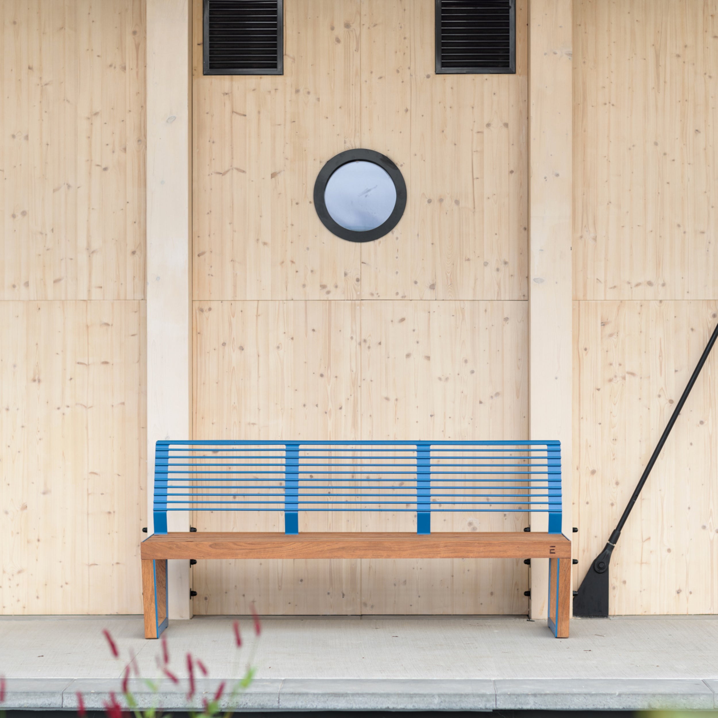 The Barka Public Bench by Egoe, made of Jatoba wood and featuring a blue metal backrest with zinc and powder coating, stands against a light wood-paneled wall. A round window is above it, flanked by two small rectangular vents near the ceiling. Red flowers slightly blur the foreground.