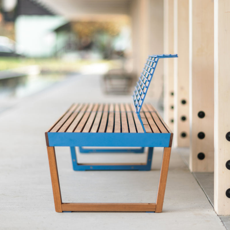 The Barka Public Bench by Egoe, with its blue metal frame and jatoba wood slats, stands on a concrete walkway. Its minimalist design features a zinc and powder-coated finish, contrasting beautifully against the blurred scenery of a covered outdoor area.