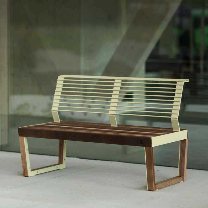 The Barka Public Bench by Egoe, featuring a Jatoba wood seat and a metal slatted backrest with zinc and powder coating, is elegantly placed on concrete in front of a large glass wall, beautifully reflecting an abstract interior as a stylish street furniture piece.