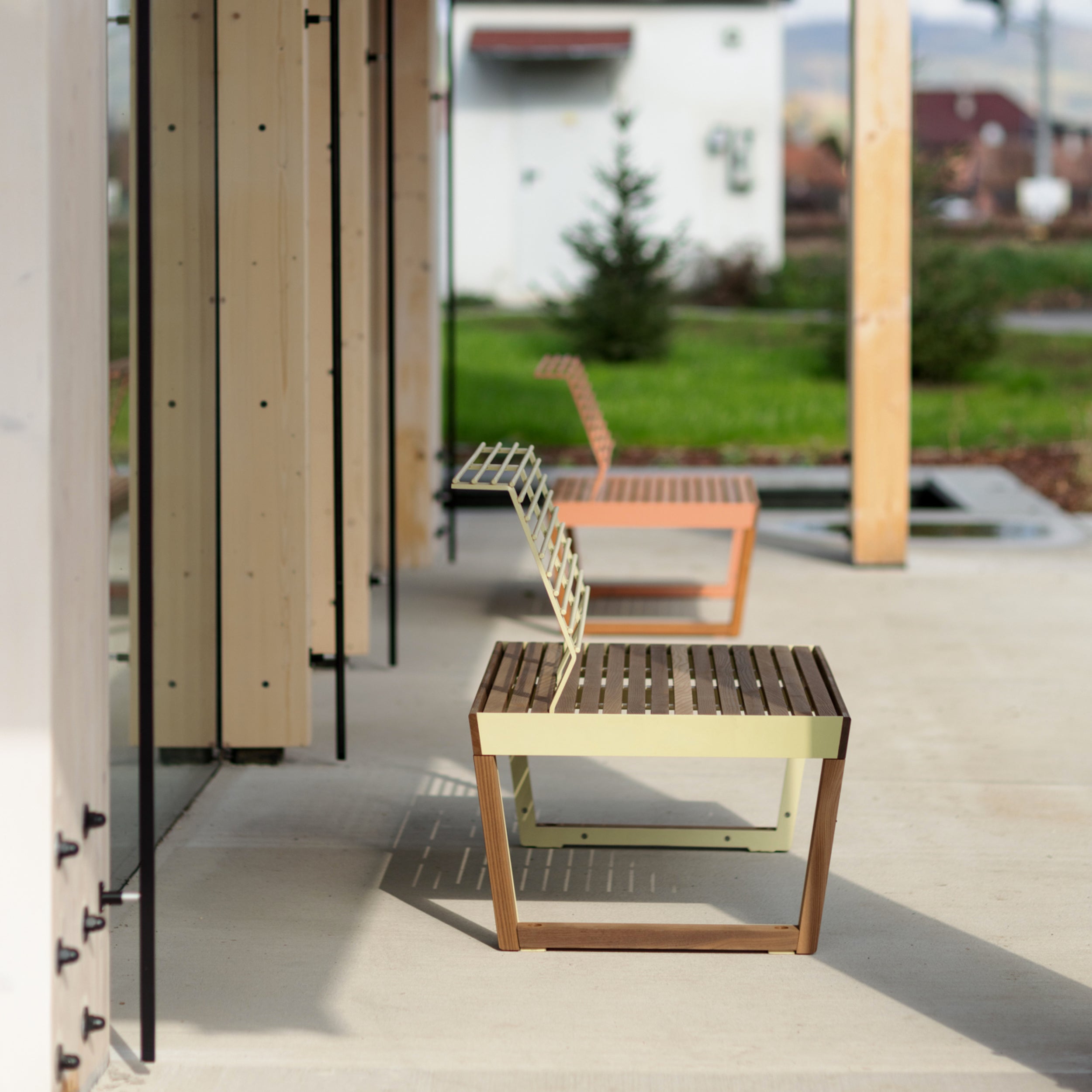 Two Barka Public Chairs by Egoé, with metal grid backrests, sit on a patio. The front chair is light green and the rear one is pink. Nearby, a small tree and buildings complement the grassy area under a clear sky.
