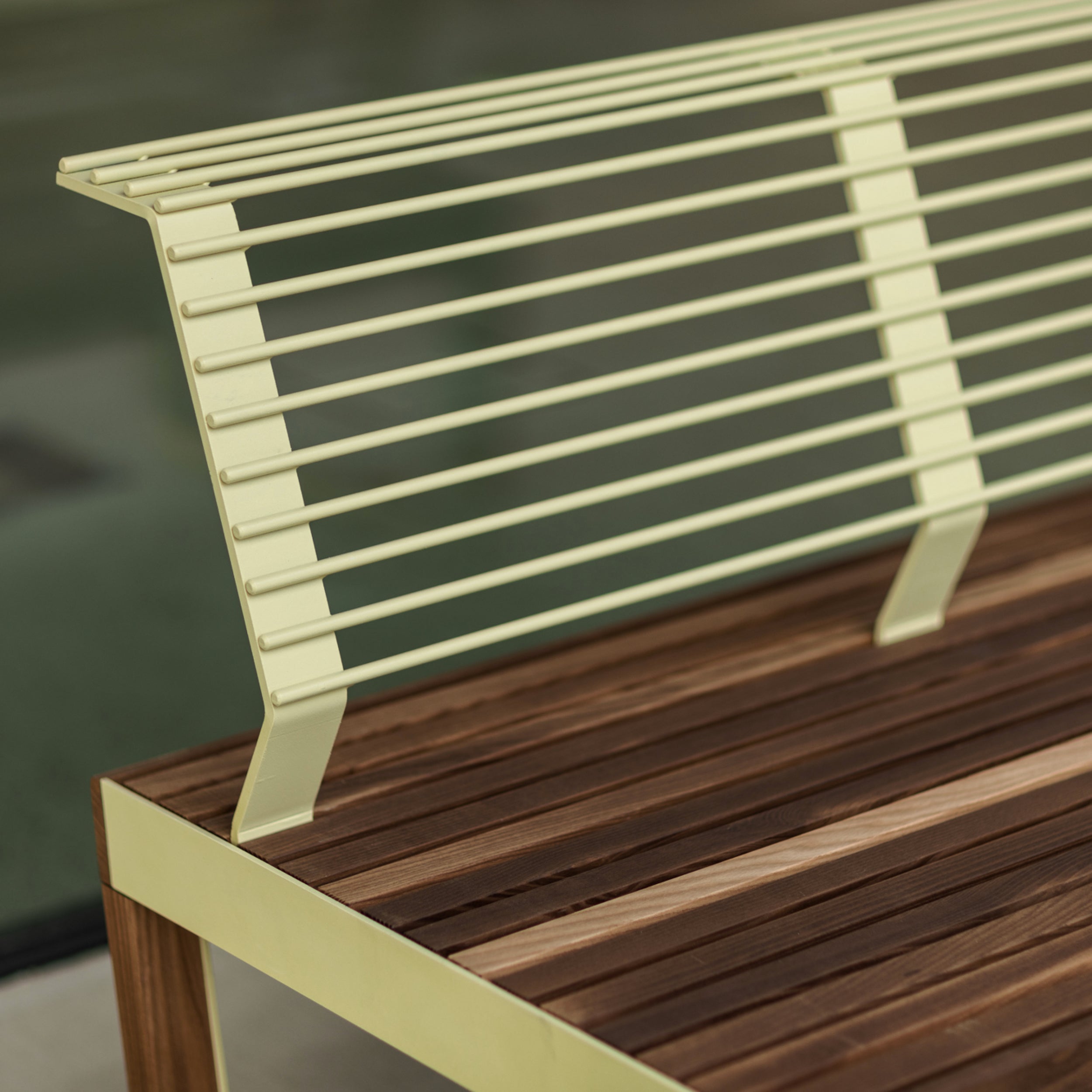 The Barka Public Bench by Egoe is a modern piece of street furniture featuring a Jatoba wood seat, slatted metal backrest, and finished with zinc and powder coating. It beautifully blends natural and industrial elements against a blurred background.