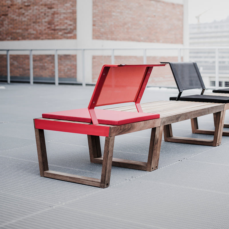 Part of the Barka collection by Egoe, this modern outdoor bench features red and black seats on a rooftop. It blends wood and metal elements, boasting a red seat partially elevated as a backrest against brick walls and metal railings.
