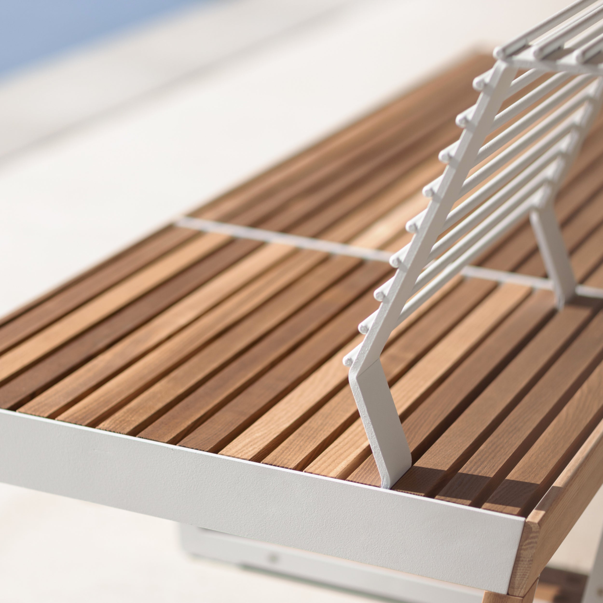 Close-up of the Barka Public Chair, an Egoé creation with tropical Jatoba wood and a sleek white metal frame. Sunlight highlights its geometric design as shadows play across the slatted panels, set against the softly blurred charm of street furniture in an outdoor scene.