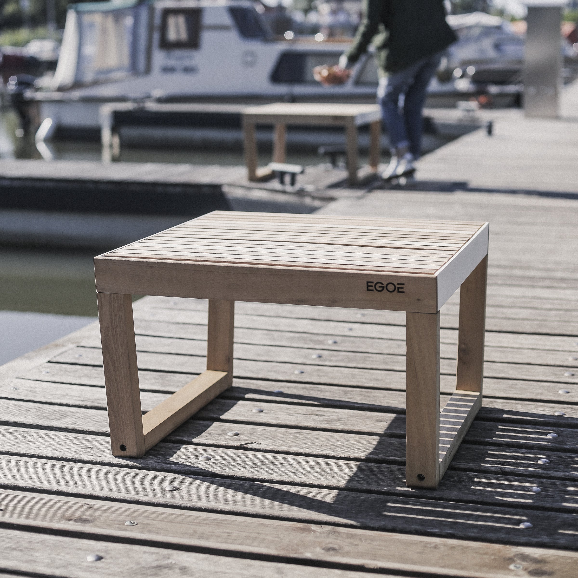 An Egoe Barka Stool adorns the dock, showcasing outstanding outdoor furniture. In the background, a person is near boats moored on serene waters under a clear sky, elevating the relaxed, sunny ambiance.