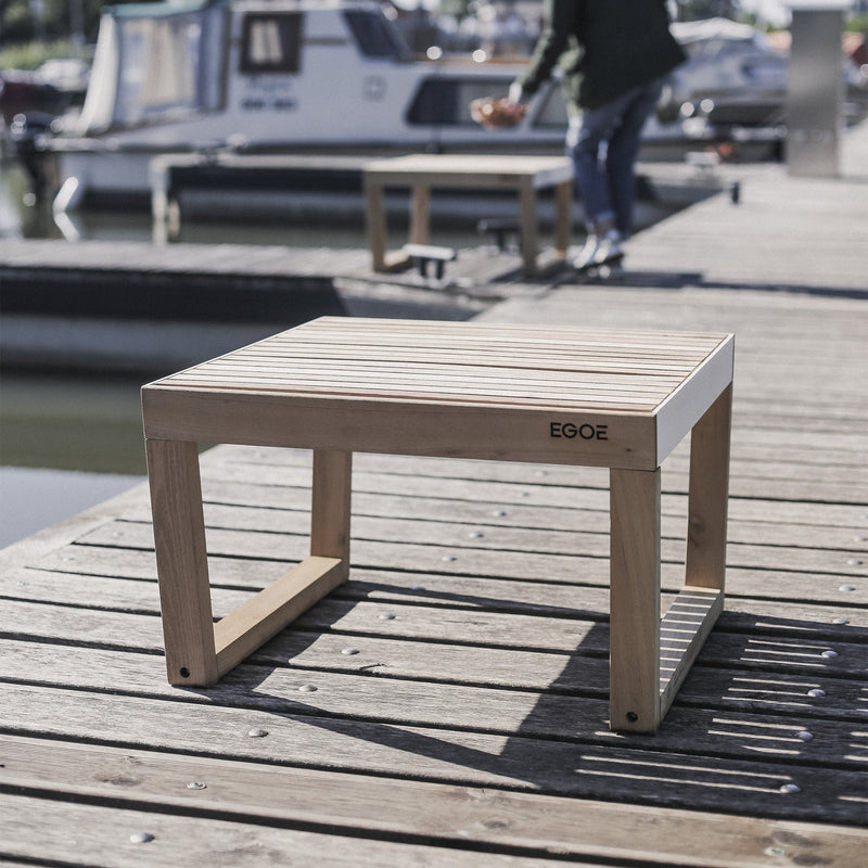 An Egoe Barka Stool adorns the dock, showcasing outstanding outdoor furniture. In the background, a person is near boats moored on serene waters under a clear sky, elevating the relaxed, sunny ambiance.
