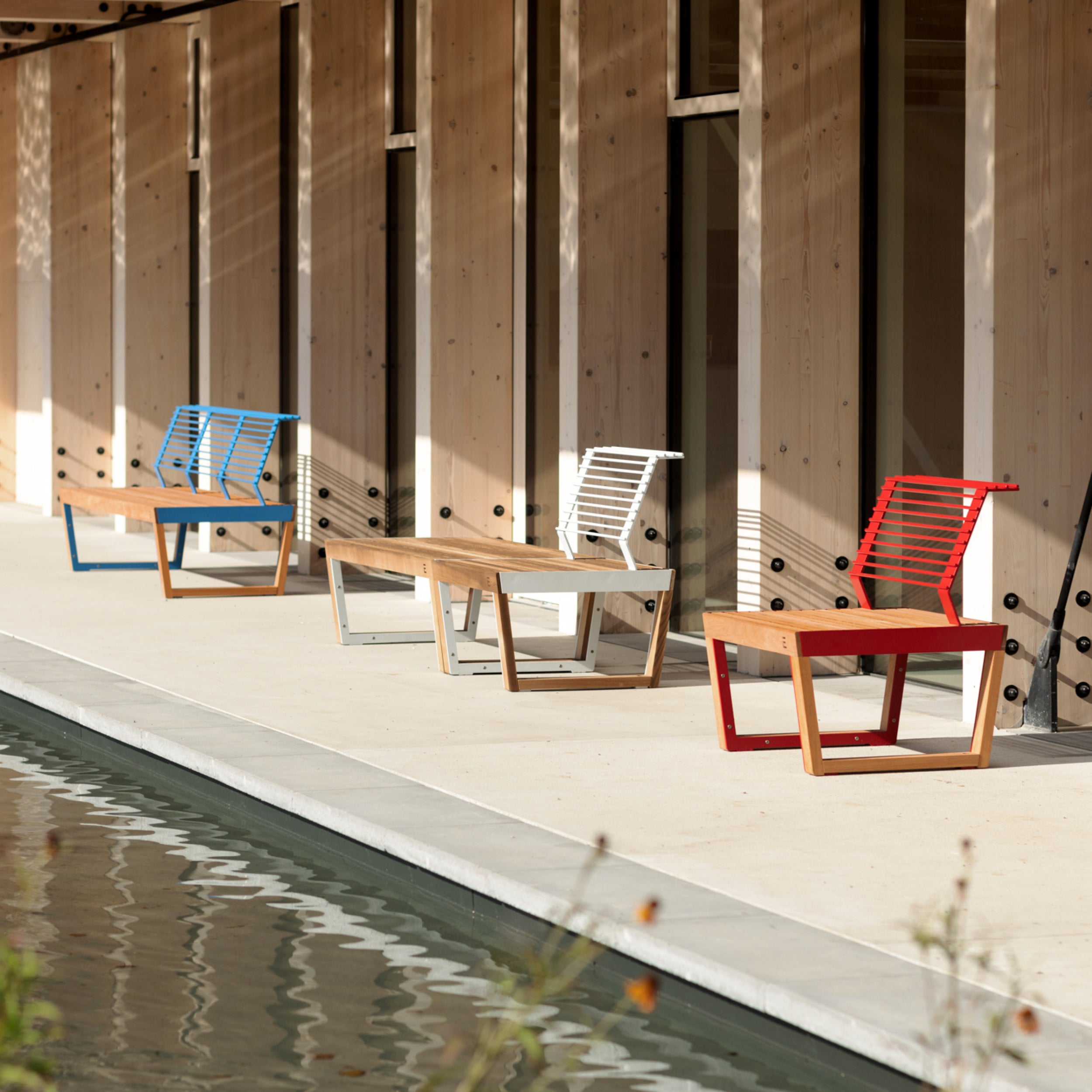 Four Barka Public Chairs by Egoe in blue, white, gray, and red with matching tables line a wooden buildings exterior beside a reflecting pool. Crafted from tropical Jatoba wood, the setting is calm, enhanced by soft light creating a relaxing ambiance.