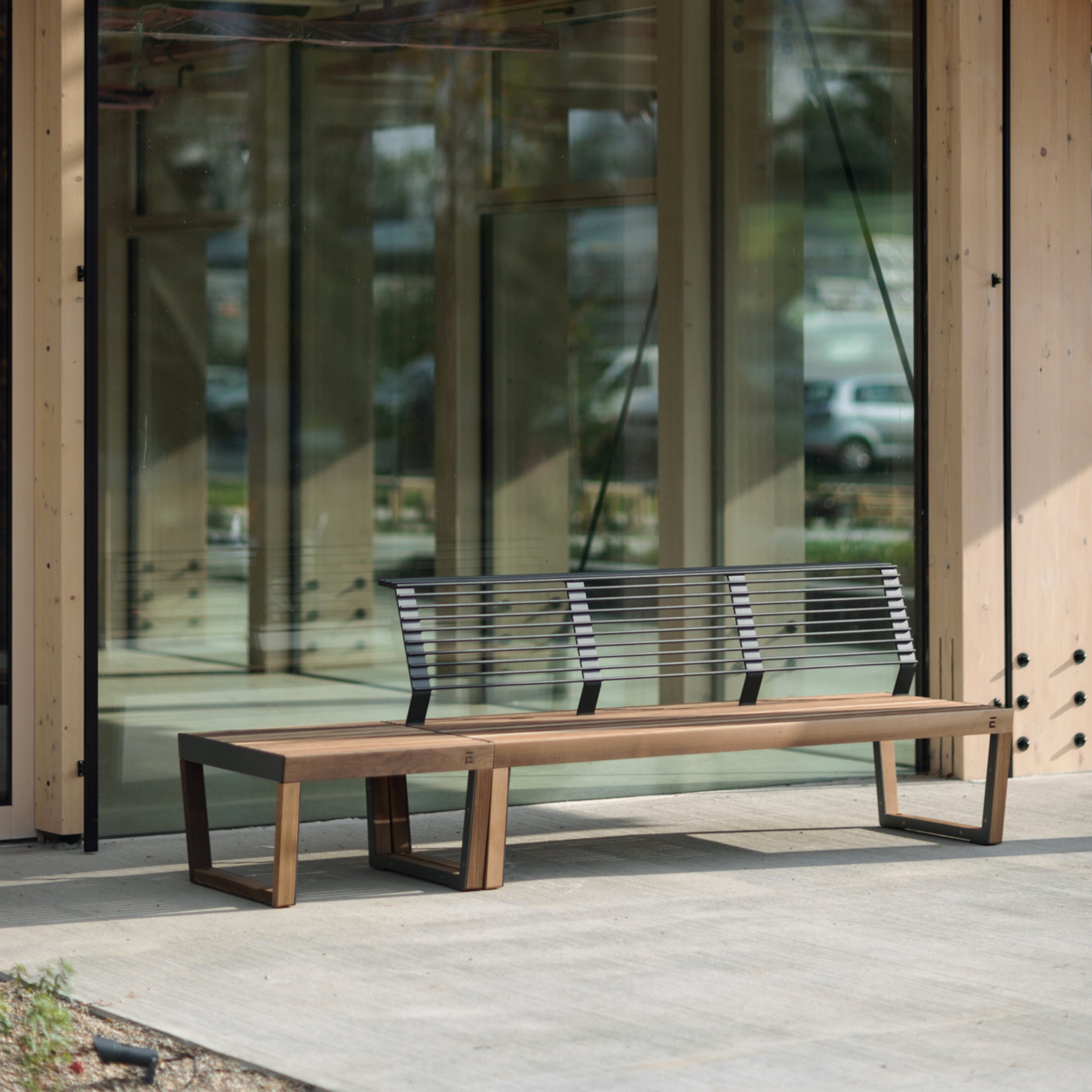 The Barka Public Stool by Egoe, crafted from corrosion-resistant materials and tropical Jatoba wood, is placed on a concrete patio. Positioned in front of a glass-windowed building, it reflects passing cars and bathes in sunlight, enhancing its relaxing charm as street furniture.