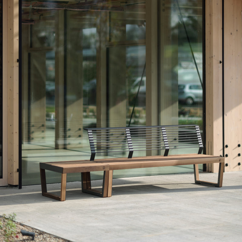 The Barka Public Stool by Egoe, crafted from corrosion-resistant materials and tropical Jatoba wood, is placed on a concrete patio. Positioned in front of a glass-windowed building, it reflects passing cars and bathes in sunlight, enhancing its relaxing charm as street furniture.