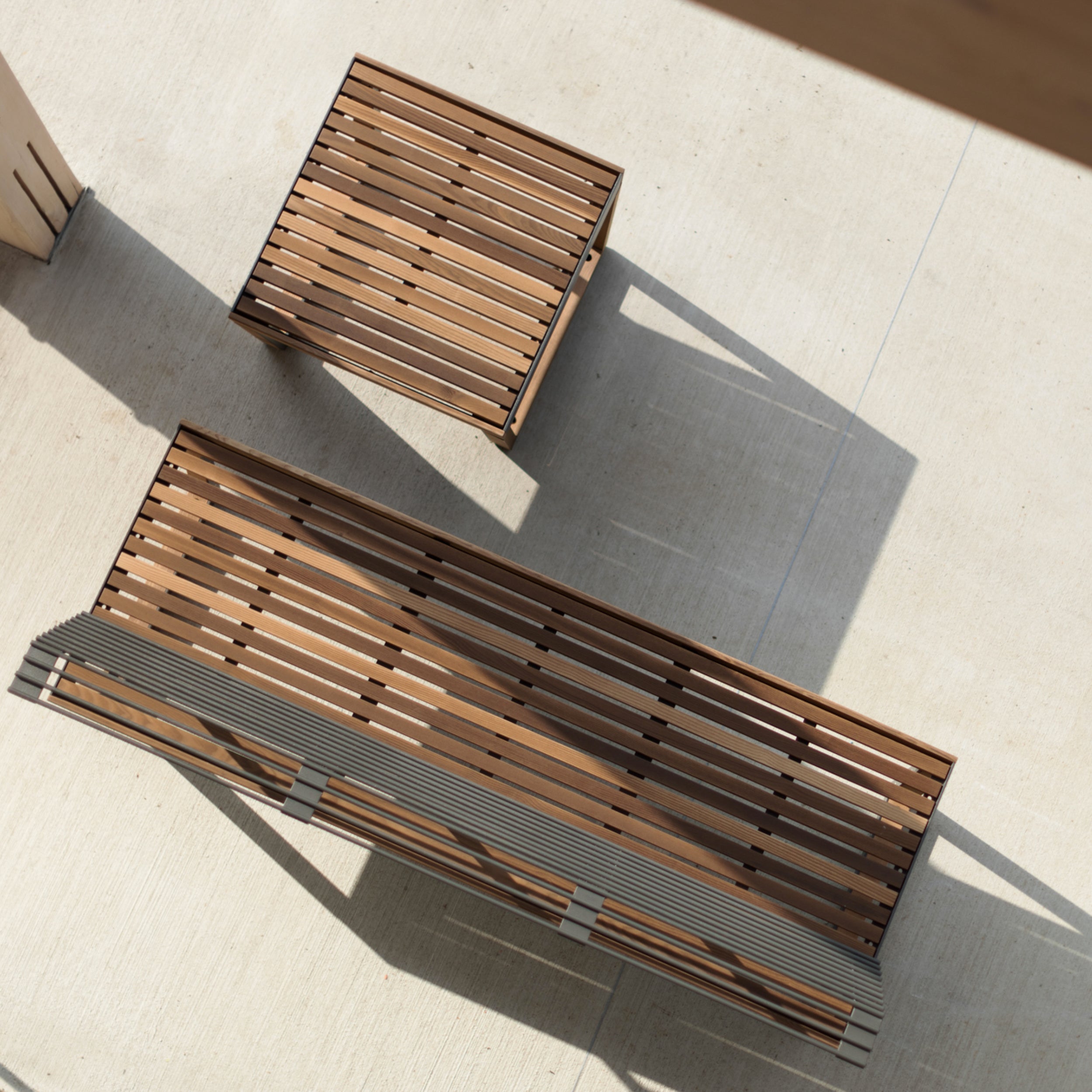 Aerial view of street furniture on concrete: Egoes Barka Public Bench, crafted from Jatoba wood with a slatted design, pairs with a small square table. The bench casts crisp shadows in the sunlight.