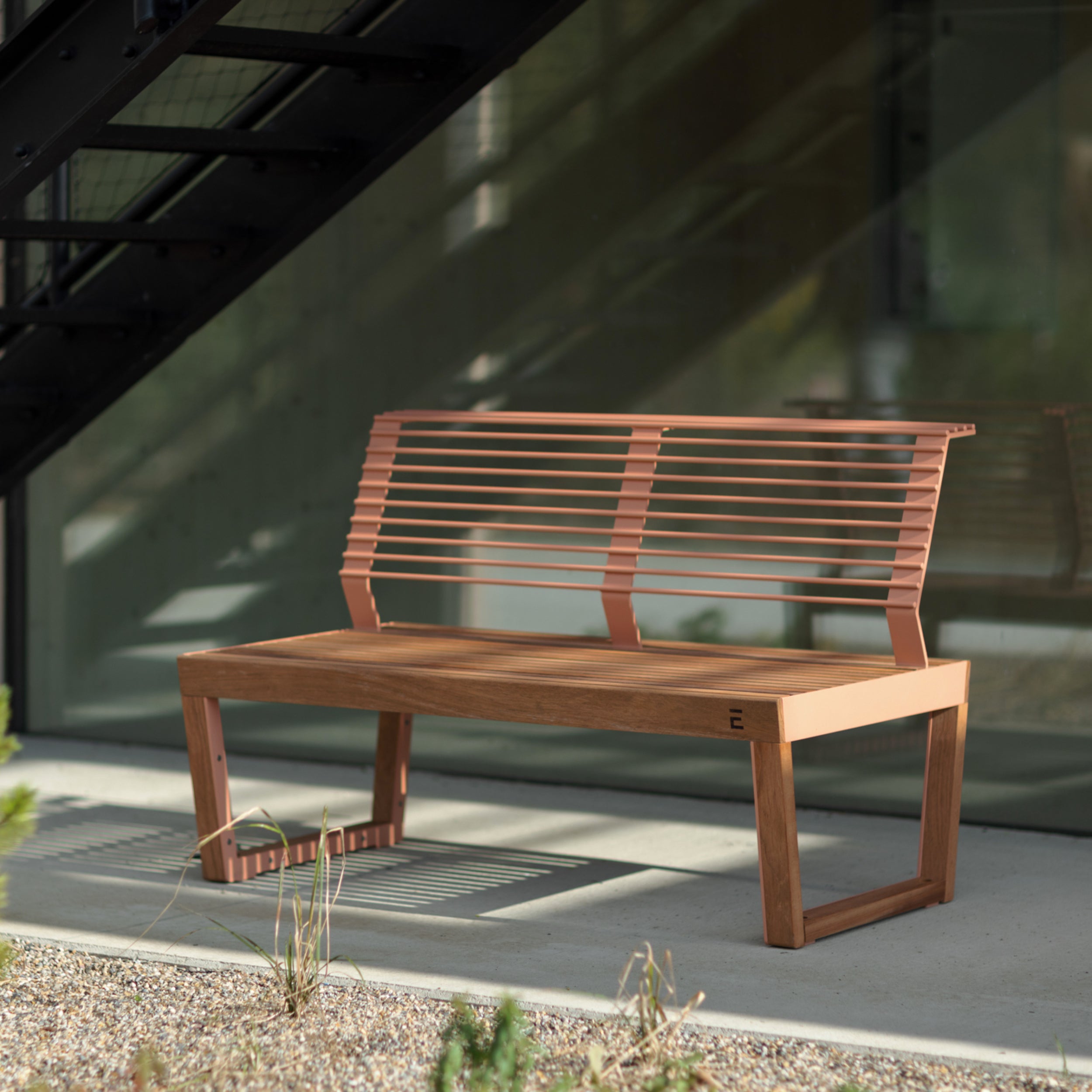 The Barka Public Bench by Egoe, crafted from Jatoba wood with a slatted backrest, sits on a concrete patio under a metal staircase. A glass wall in the background reflects greenery while sunlight casts shadows on the ground.