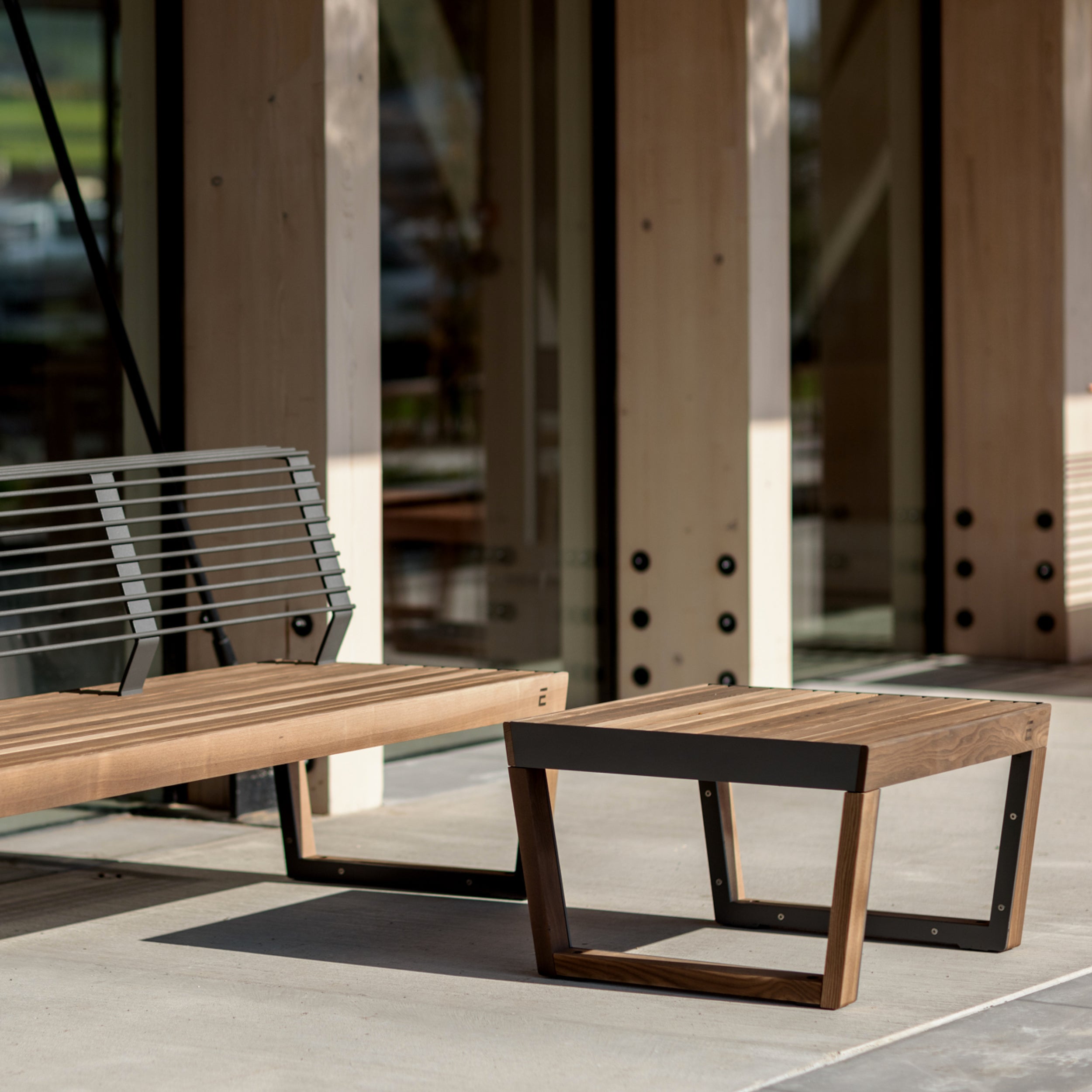 A Barka Public Stool from Egoe, made of tropical Jatoba wood, complements a modern patio with glass and wooden walls. Sunlight casts warm shadows on the concrete floor, crafting an inviting space ideal for enjoying this durable street furniture.