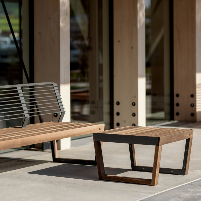 A Barka Public Stool from Egoe, made of tropical Jatoba wood, complements a modern patio with glass and wooden walls. Sunlight casts warm shadows on the concrete floor, crafting an inviting space ideal for enjoying this durable street furniture.