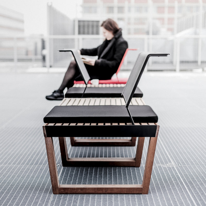 A modern outdoor setting showcases Barka collection wooden slat benches by Egoe. Nearby, a sleek black Barka Chair stands out. In the background, a blurred reader relaxes amidst urban structures, enjoying the versatile design.