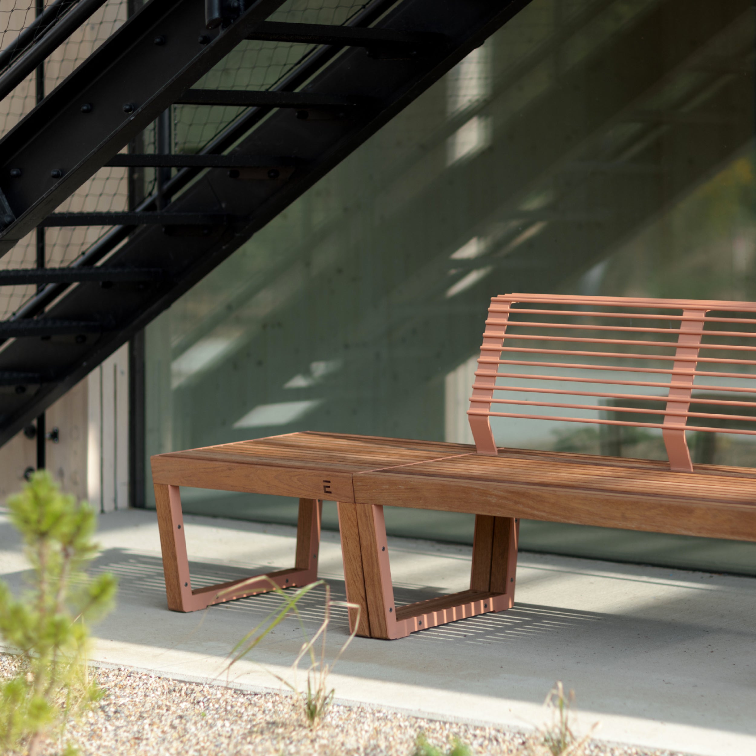 The Barka Public Stool by Egoe, crafted from tropical Jatoba wood with a slatted backrest, sits on a concrete surface. Its positioned under a black metal staircase. Tall glass windows reflecting greenery create a serene outdoor setting, making it ideal for stylish street furniture.