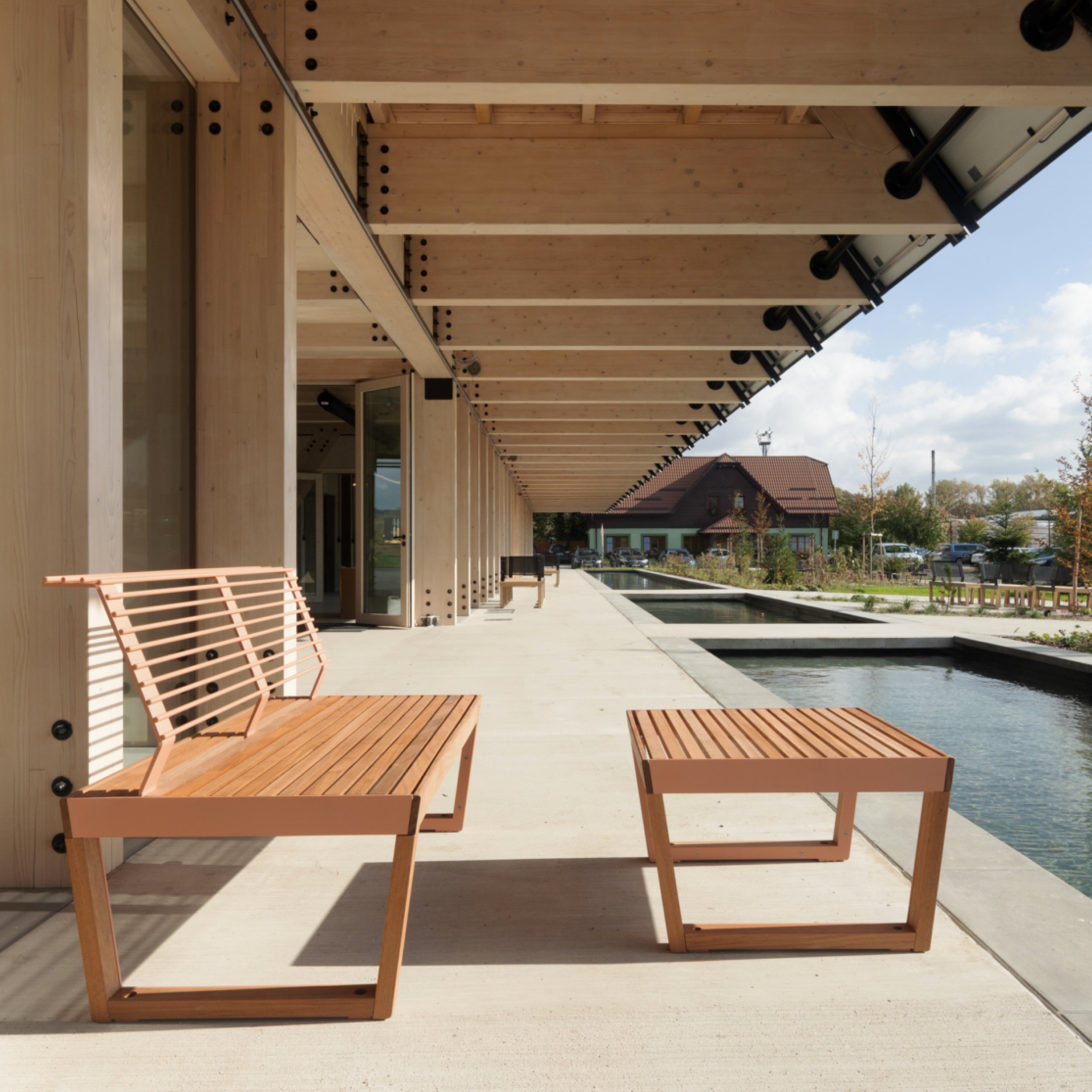The outdoor area features Barka Public Stools by Egoe under a pergola with wooden benches and tables crafted from tropical Jatoba wood. A tranquil pond lines the pathway, with lush greenery and a wooden building visible against a partly cloudy sky.